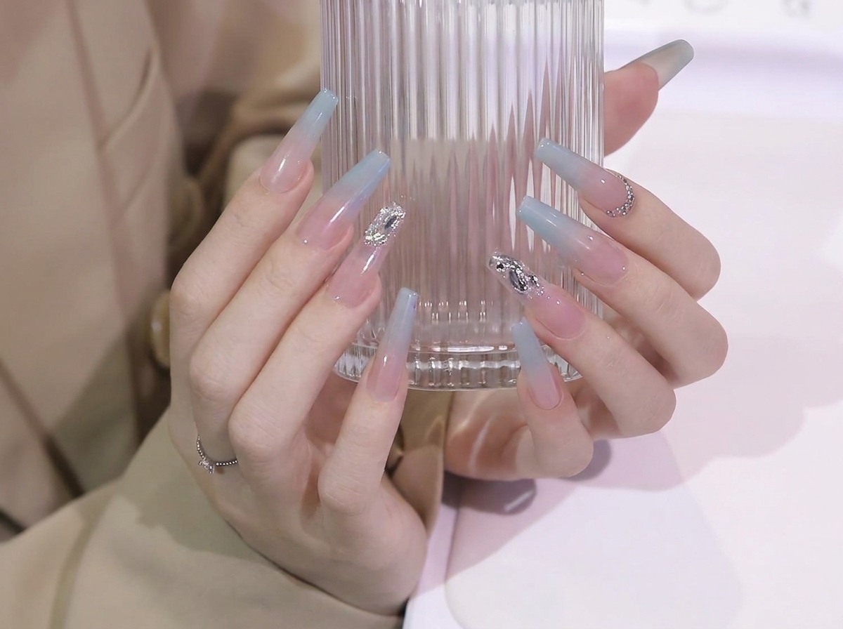 Hands adjusting grip on a glass cup to showcase the smooth blue-to-nude gradient transition on long coffin nails.