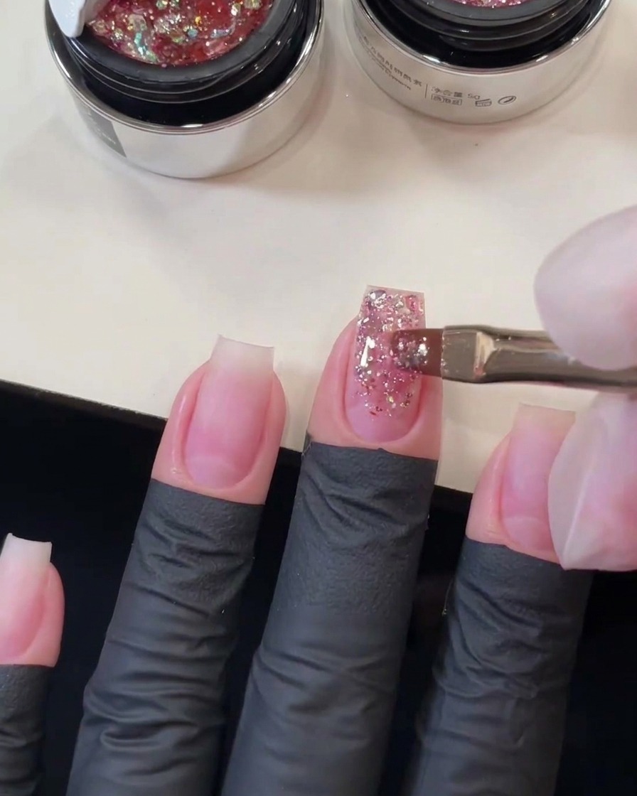 Flat gel brush sweeping pink chunky sequins down the length of the middle fingernail to form a glitter gradient.