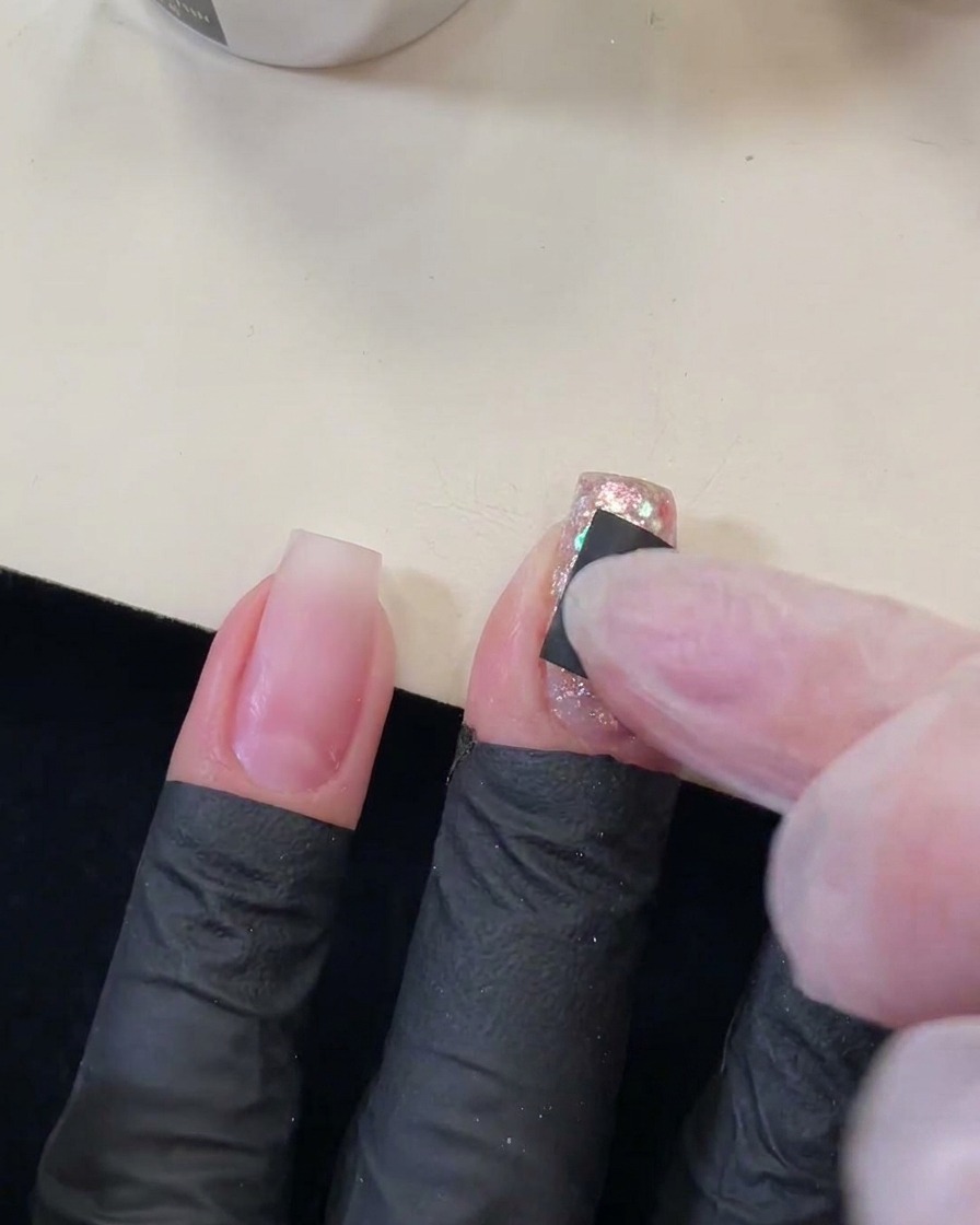 A finger pressing a small black square sticker with a star-shaped cutout onto a sparkly, sequined nail.