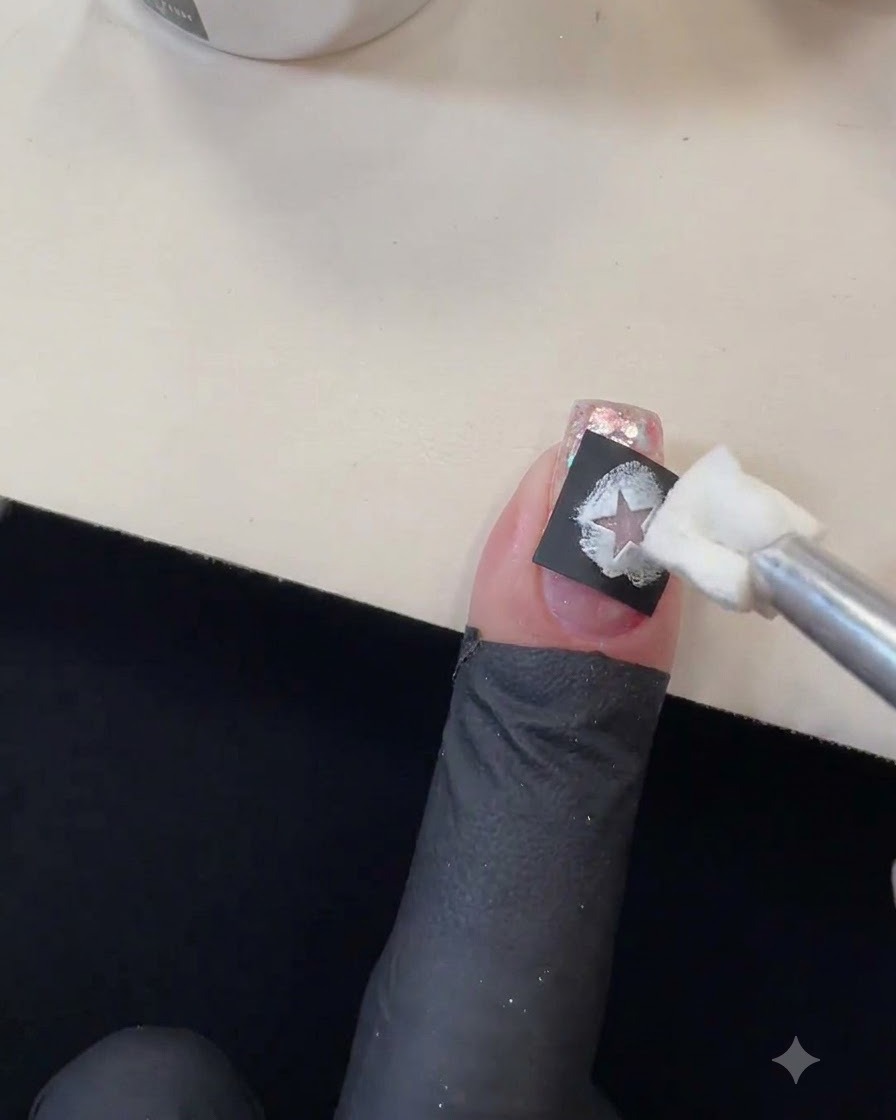 A sponge applicator dabbing white gel polish into the cutout of a black star stencil on a glittery nail.