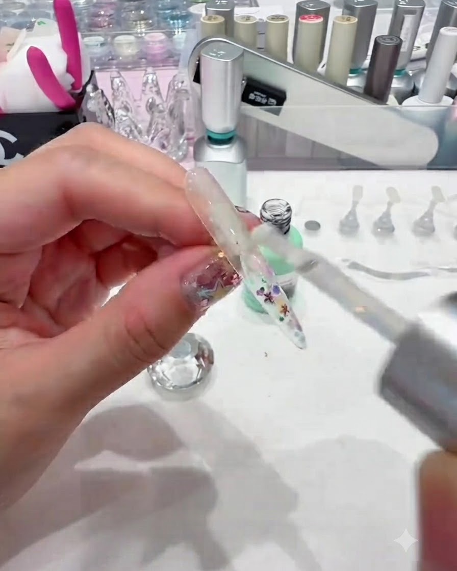 A brush applying a thick layer of clear builder gel over a sequined nail tip to encapsulate it smoothly.