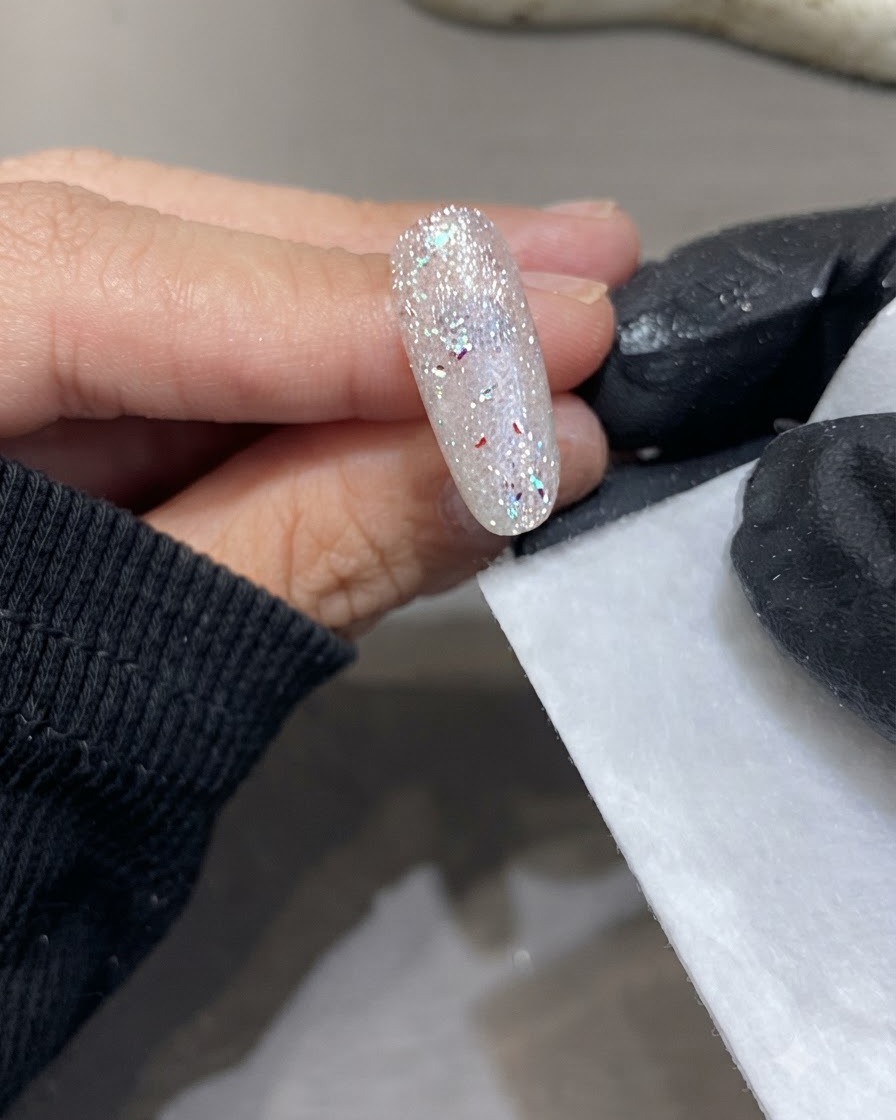 A gloved hand holding a lint-free wipe next to a cured sparkling nail surface.