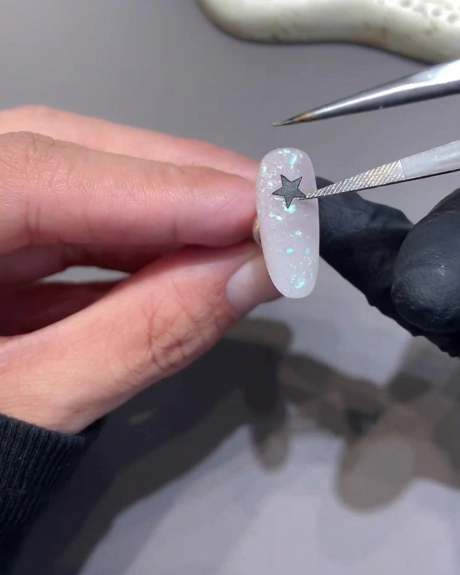 Tweezers placing a black star sticker onto a glittery pink nail surface.