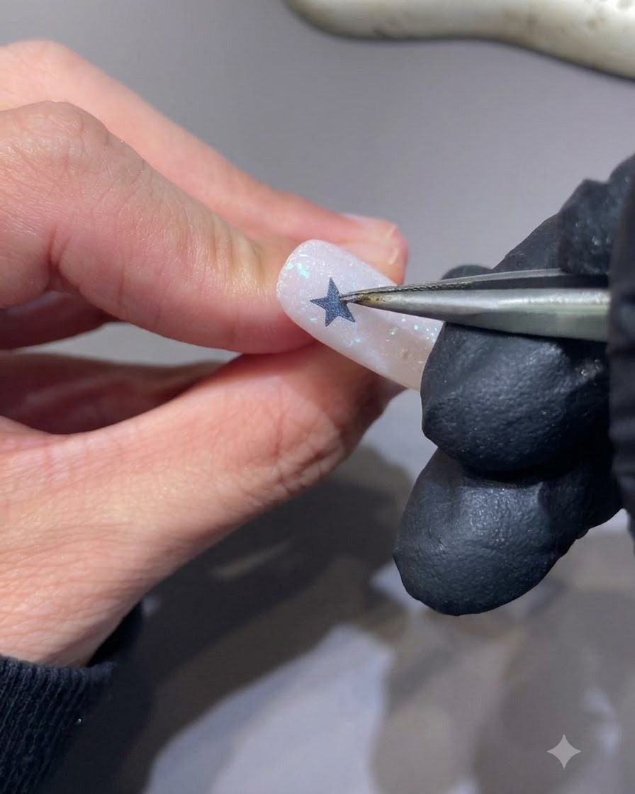 Tweezers peeling off a star sticker from a nail to reveal a hollow star silhouette.