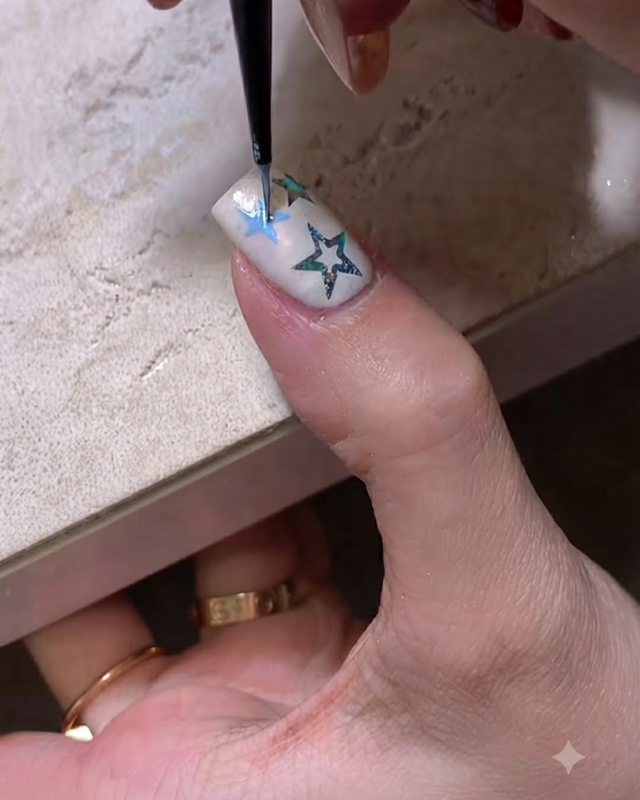 A close-up of a fine nail art brush drawing a blue star outline on a manicured nail.