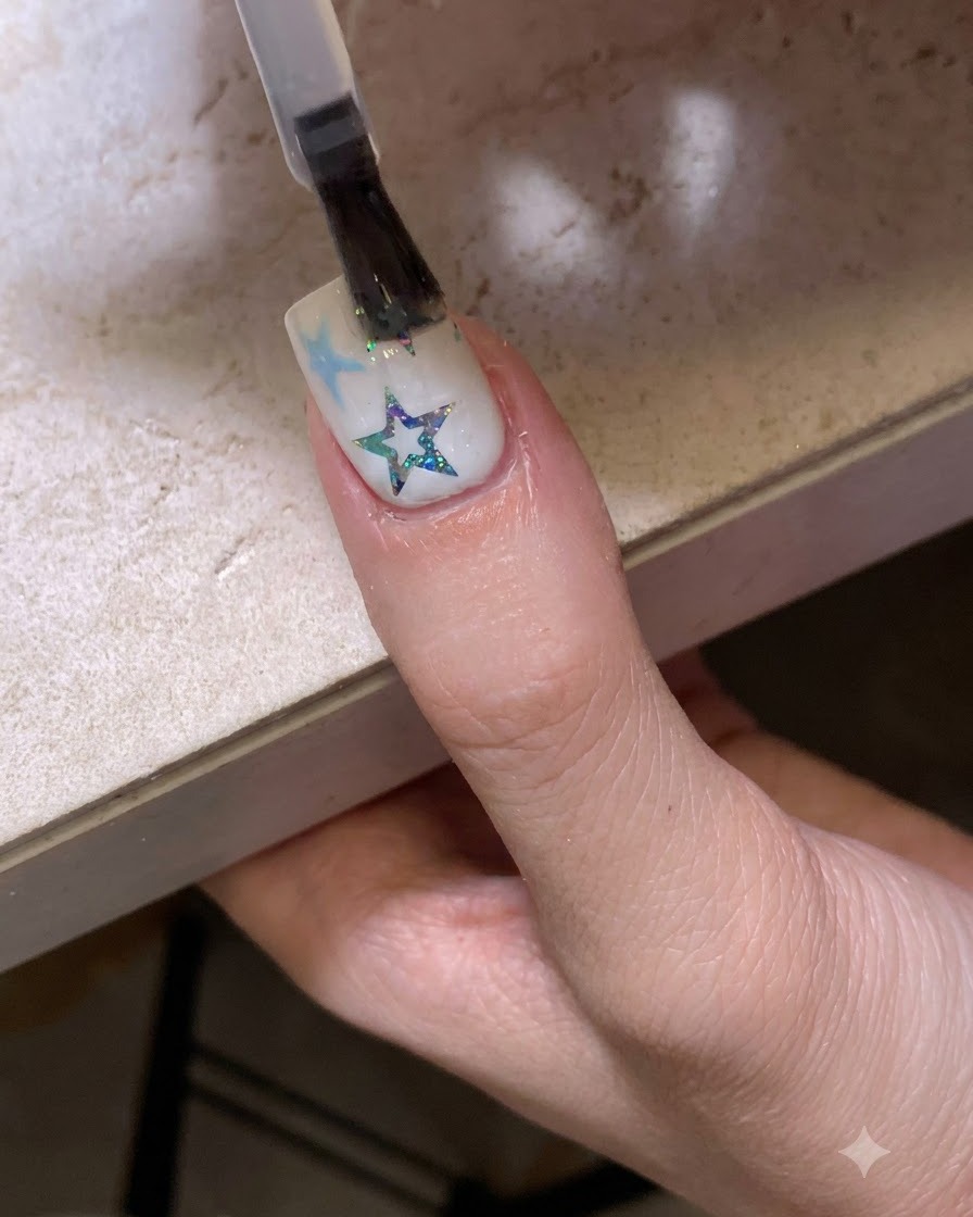A brush applying a glossy clear top coat over a nail decorated with stars and glitter.