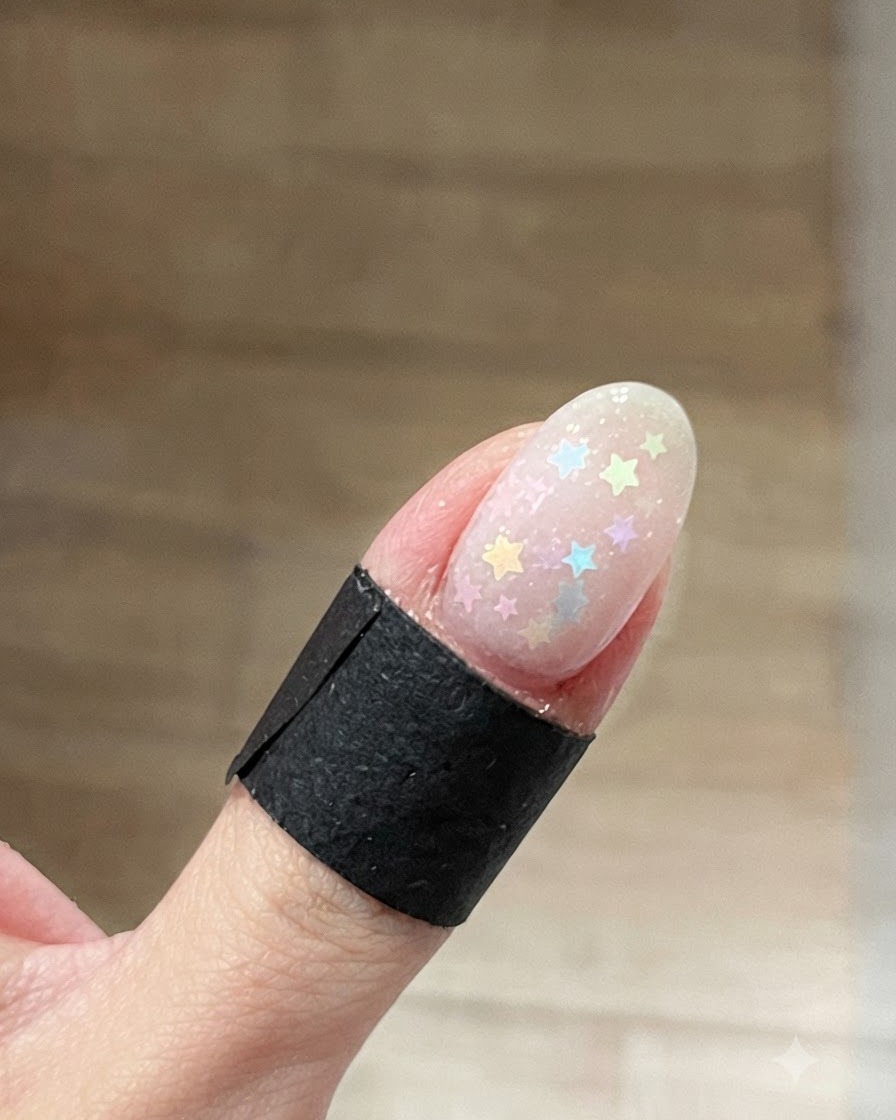 A close-up of an oval fingernail with pastel multi-colored star glitters scattered across a clear wet base gel.
