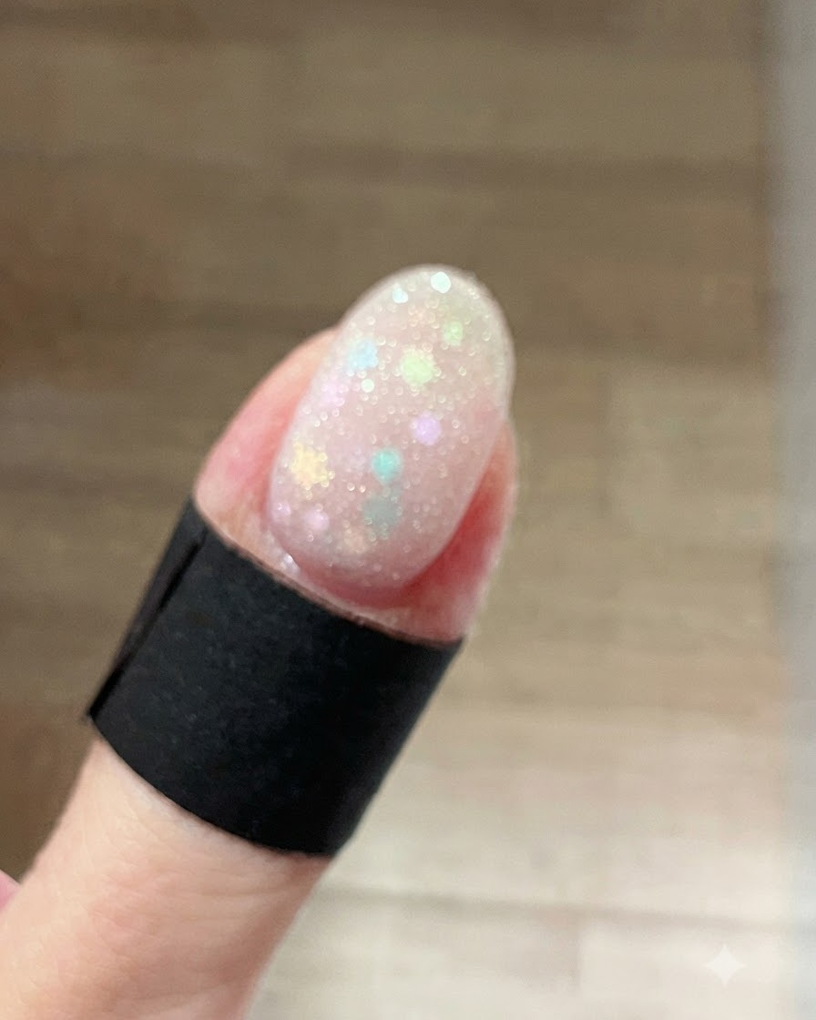 A close-up of a fingernail coated with a wet, sparkly frost white shimmer gel layered over pastel star glitters.