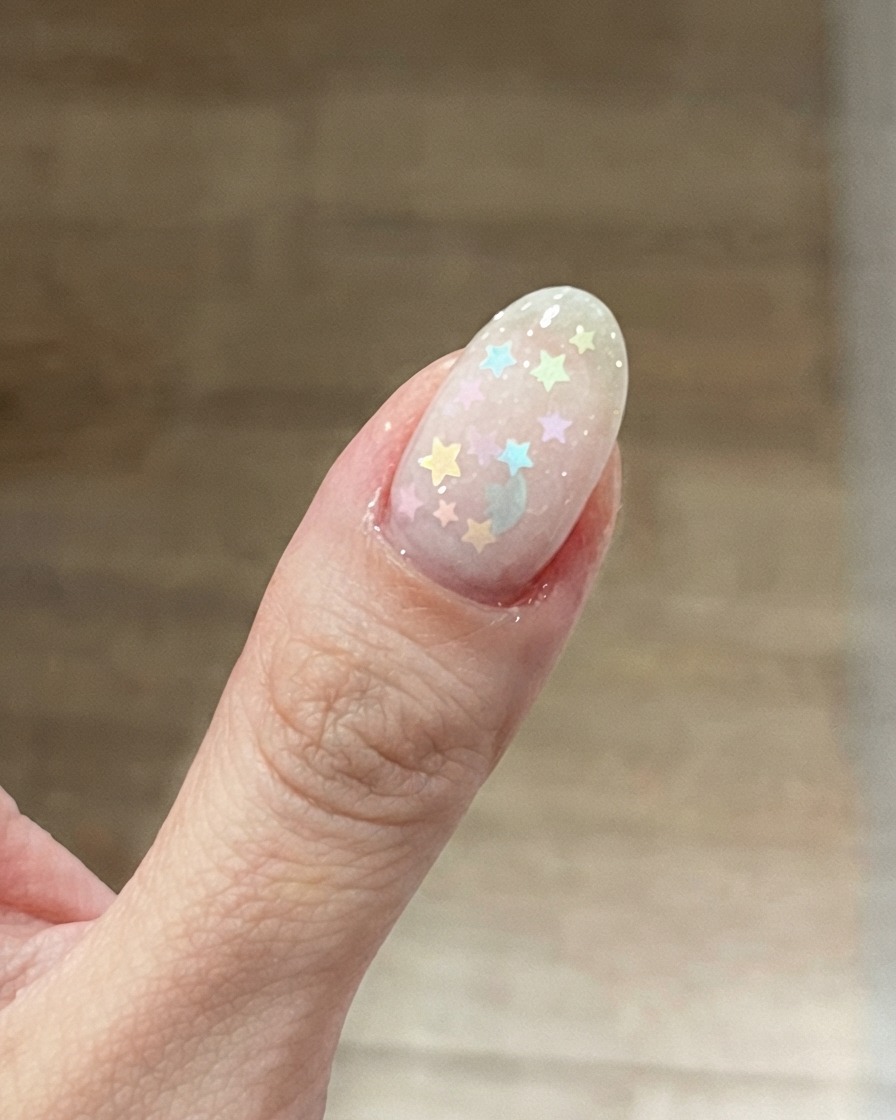 A close-up of a thumb nail featuring a milky pink base with colorful pastel star glitters placed on top.
