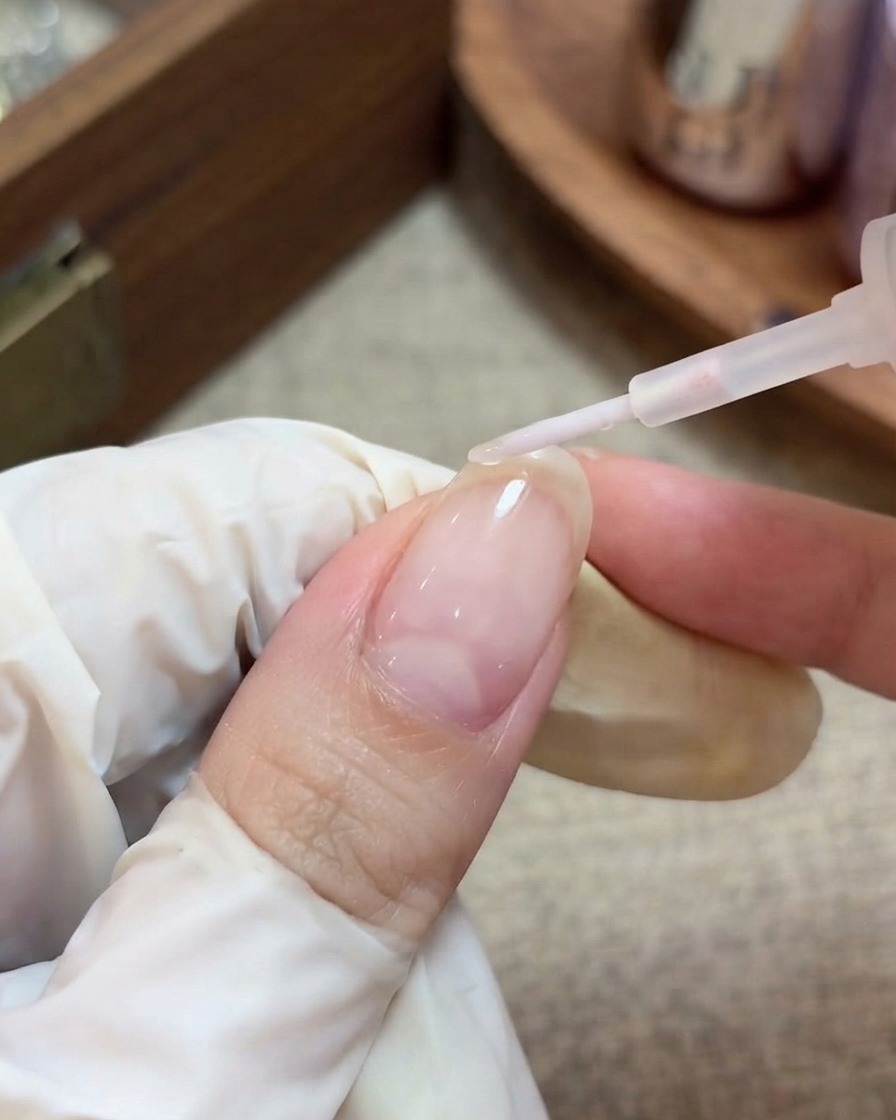 A gloved hand using a brush to apply a clear, glossy top coat over a prepped nail with encapsulated star glitters.