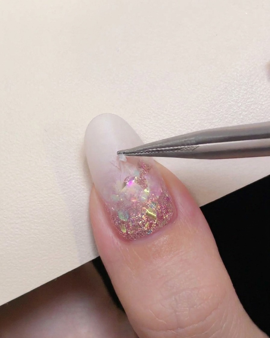 Precision tweezers carefully peeling a hollow star sticker off a milky white gradient nail to reveal pink glitter underneath.
