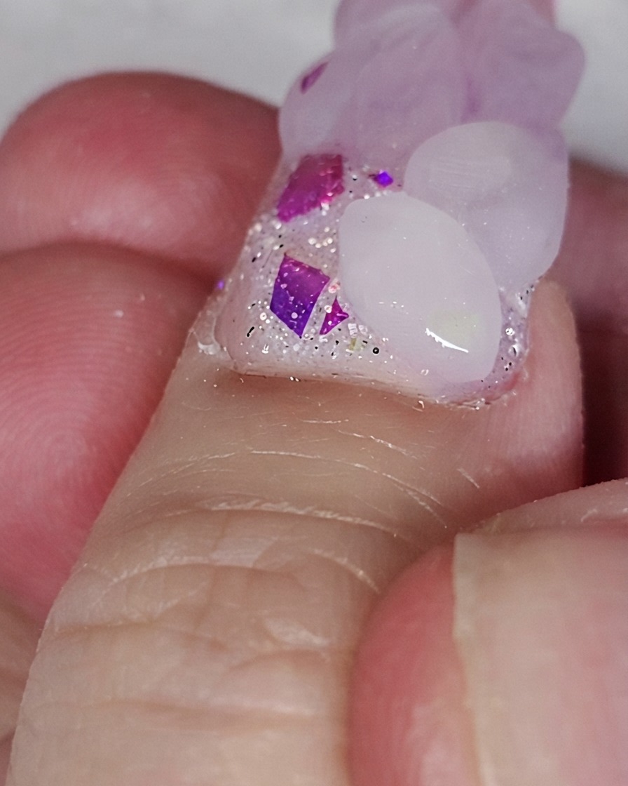 A nail brush placing a pink acrylic bead onto a glitter nail base to form a heart shape.