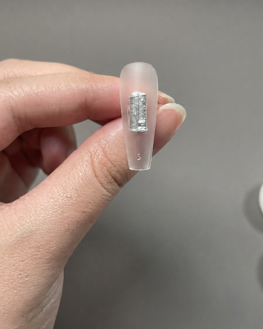 A clear nail extension piece held between fingers showing a silver or black brush stroke applied directly without a base coat.