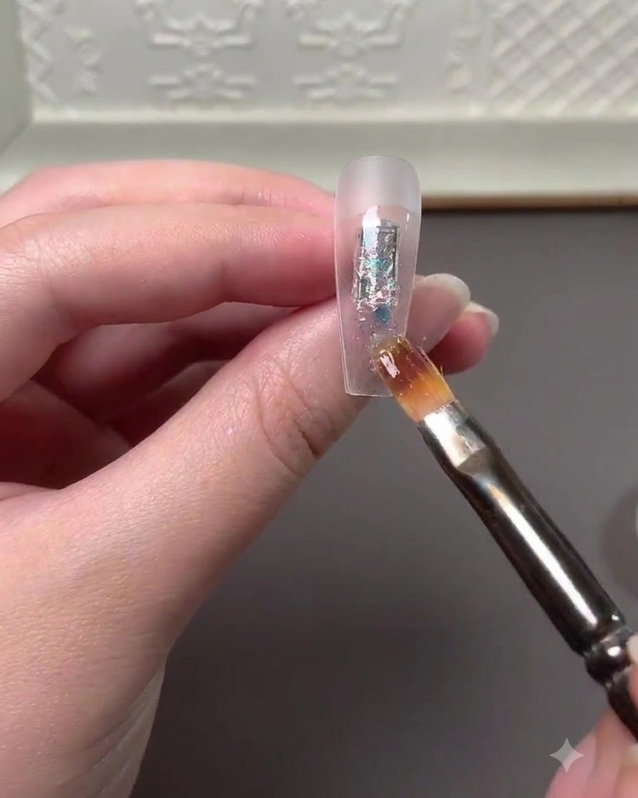 A nail art brush applying multi-colored glitter flakes and high gloss powder onto the center of a clear nail extension.
