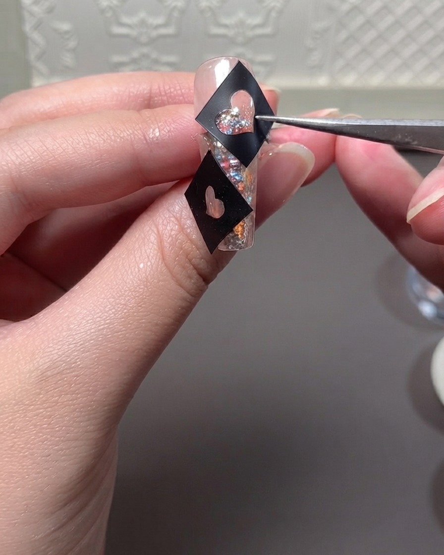 A pair of tweezers holding a black heart stencil template over a glitter-filled nail while applying gel.