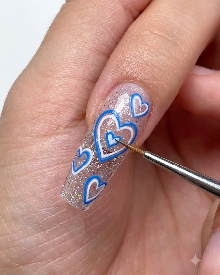 A fine detailing brush using white drawing gel to outline small dimensional hearts on a sparkly extended nail.