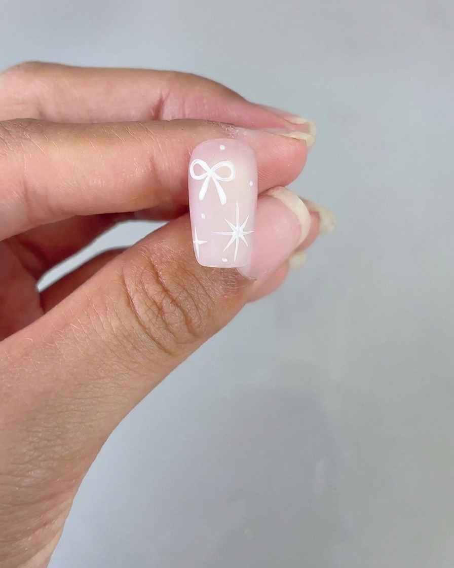 A finished nail design featuring a white bow and a four-pointed star on a pink gradient base held up for viewing.