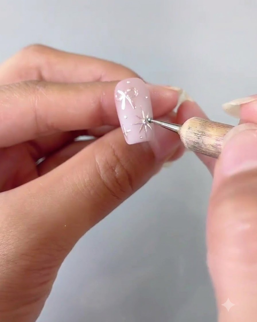 A close-up showing a dotting tool carefully placing a tiny crystal onto the center of a silver four-pointed star painted on a pink nail.