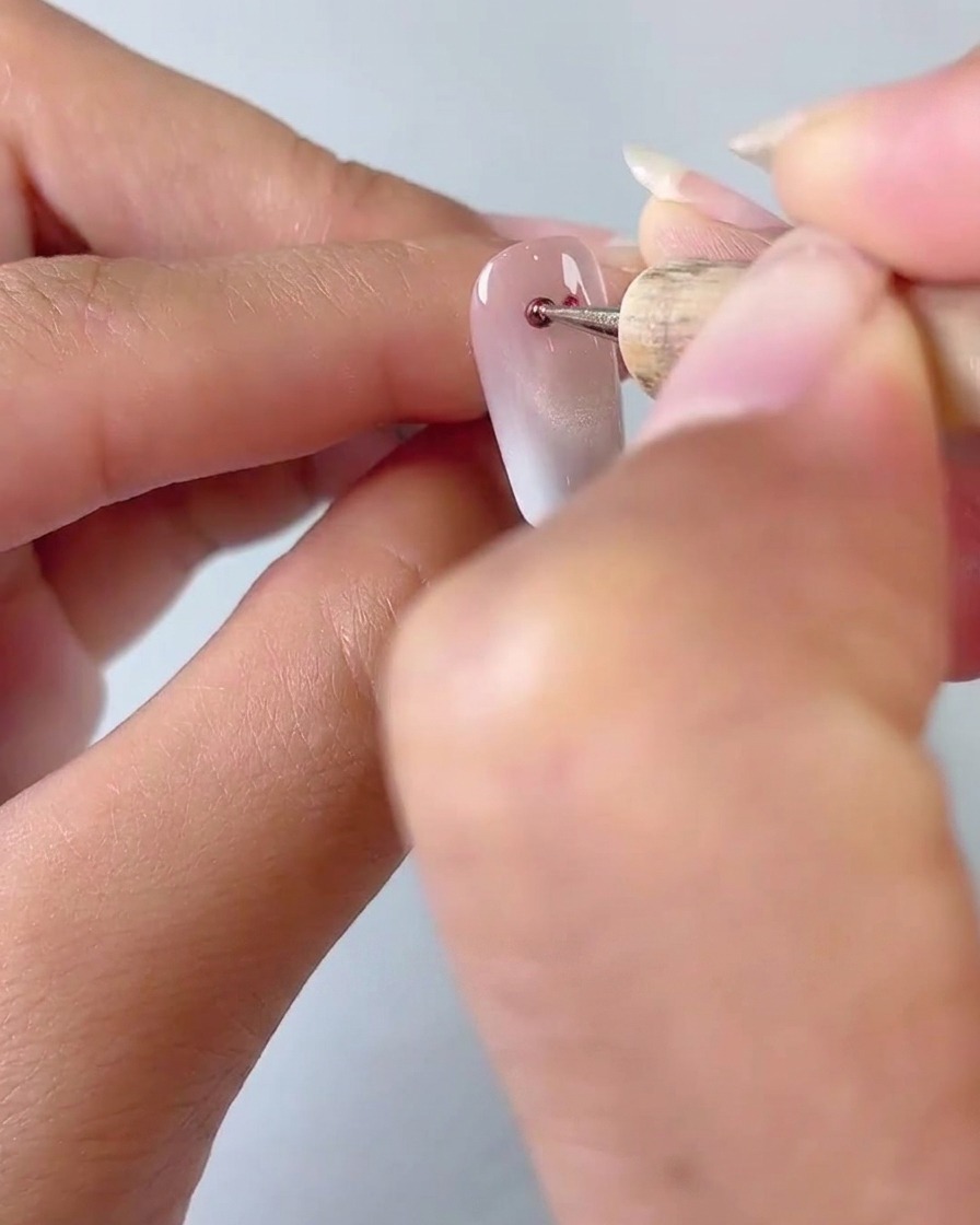 A close-up of a dotting tool applying two small dots of red cat eye gel onto a milky gradient nail base.