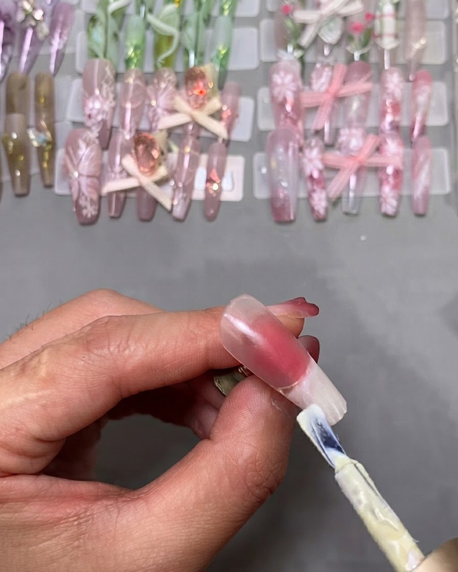 A gel polish brush applying white polish to the tip of a fake nail mounted on a stand.