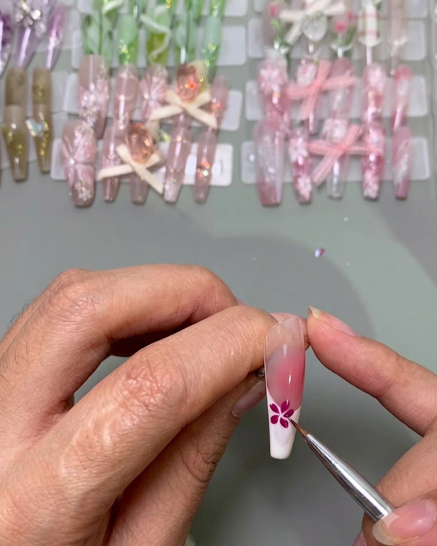 A fine brush adding blue petals next to a completed pink flower on a nail tip.