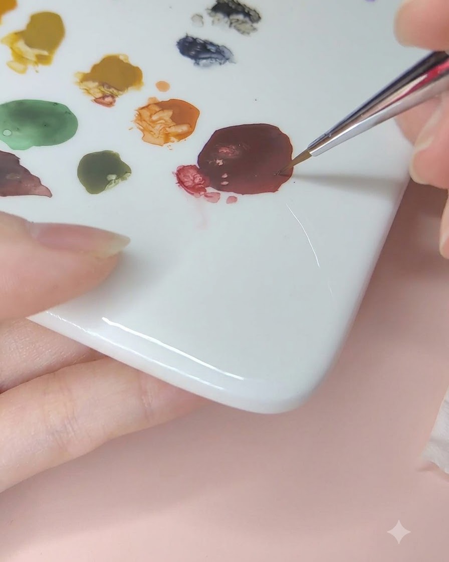 A fine nail art brush picking up dark red watercolor paint from a ceramic palette.