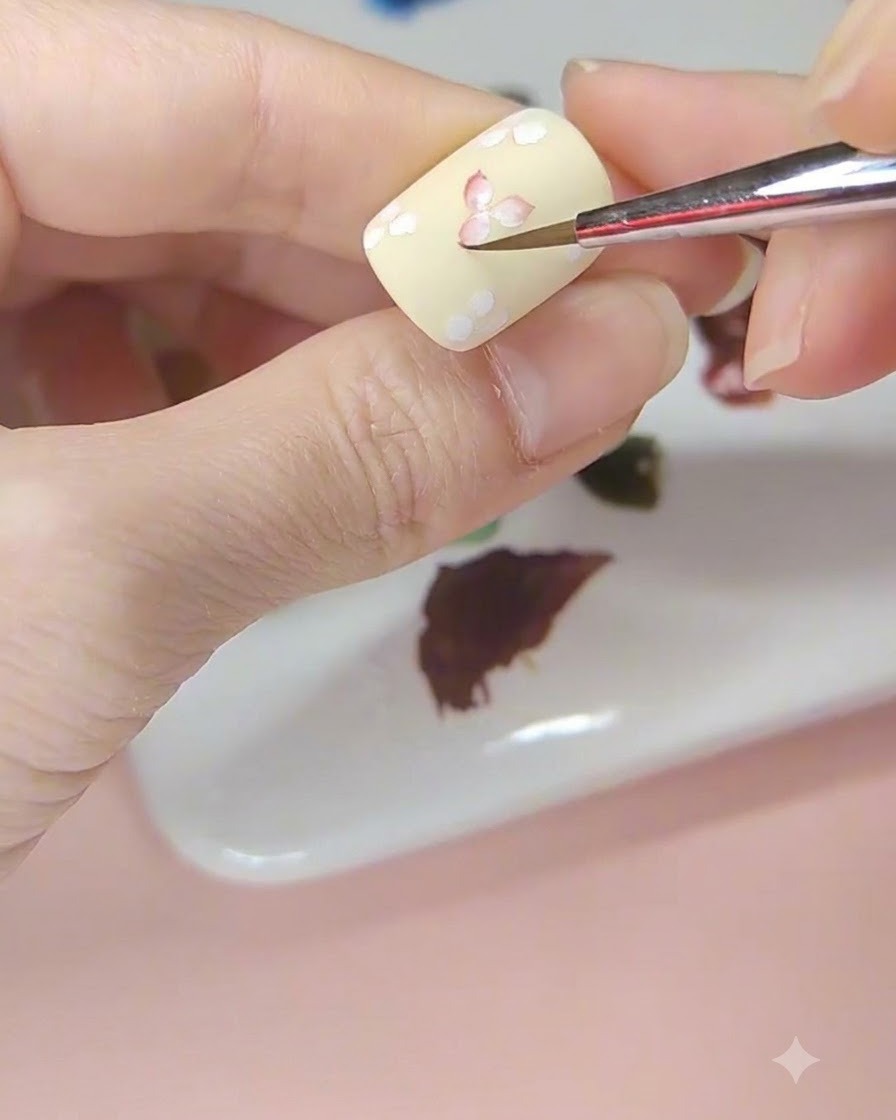 A nail art brush tinting the center of a white flower with red watercolor on a pale yellow nail.