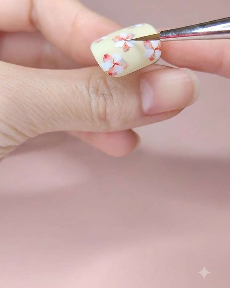 A fine nail art brush adding a touch of red watercolor pigment to the center of a white painted flower on a pale yellow nail.