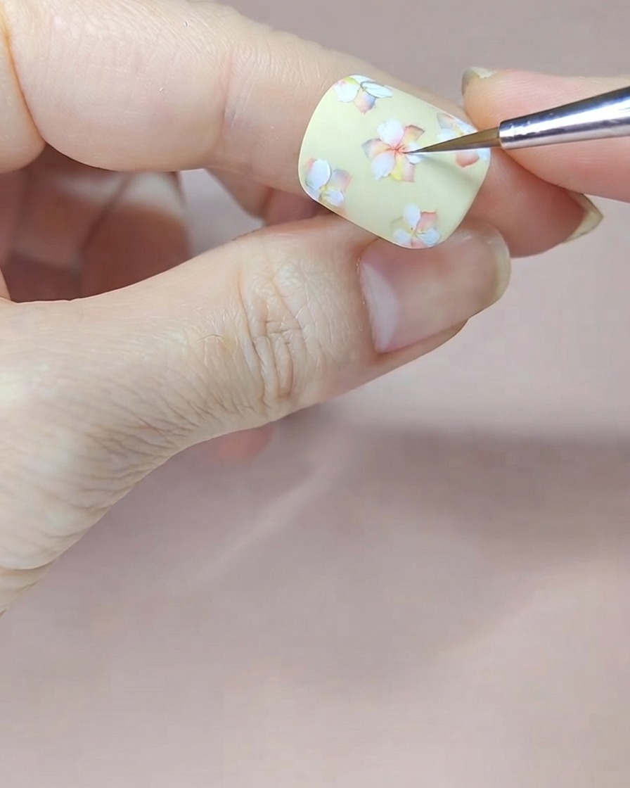 A fine brush applying red watercolor tint to the centers of white flowers on a pale yellow nail.