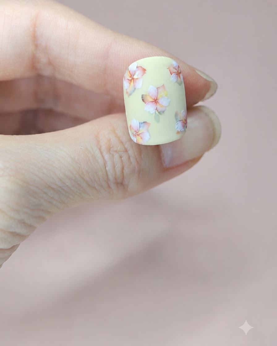 A close-up of a pale yellow nail showing a tiny green watercolor leaf painted next to the middle floral cluster.