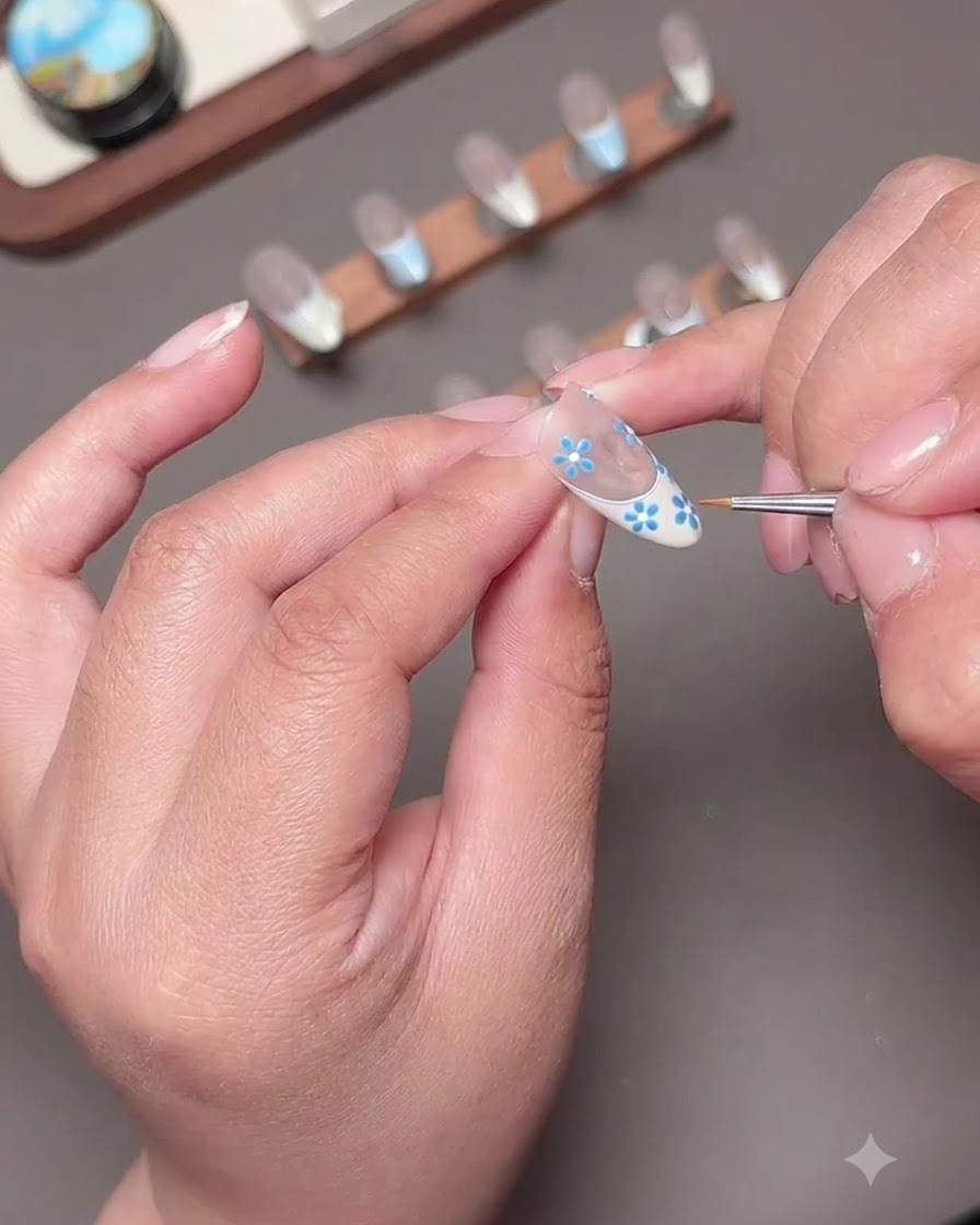 A macro shot of tiny blue petals being painted onto the nail surface with a detail brush.