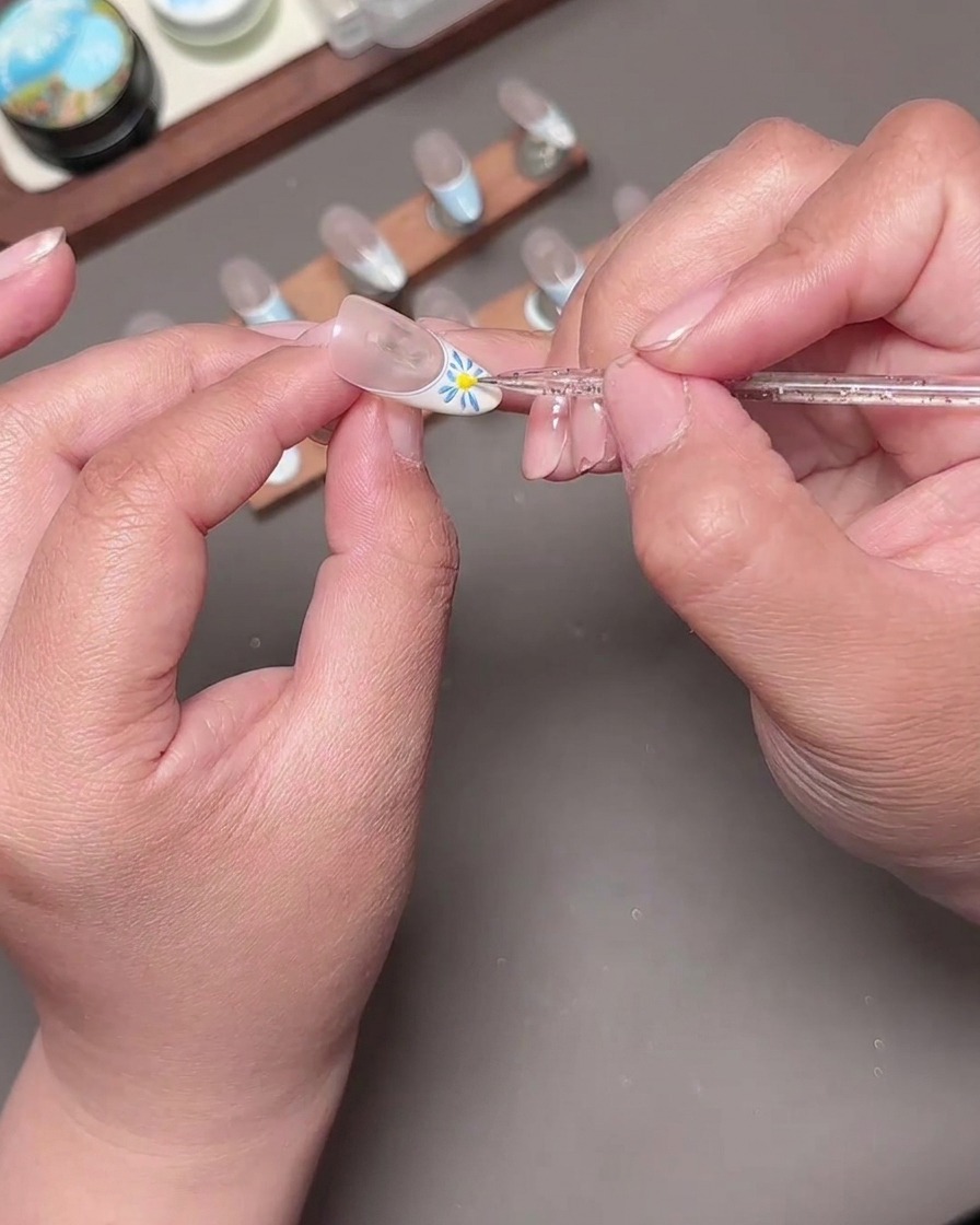 A dotting tool placing a bright yellow center dot into a blue painted flower on a French manicure nail.