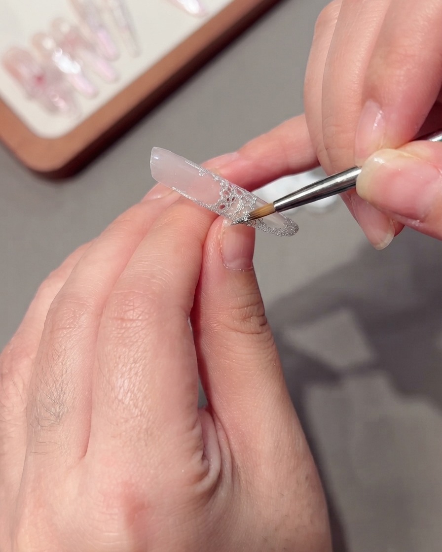A thin liner brush applying silver glitter wire-drawing gel along the edge of a lace nail design.