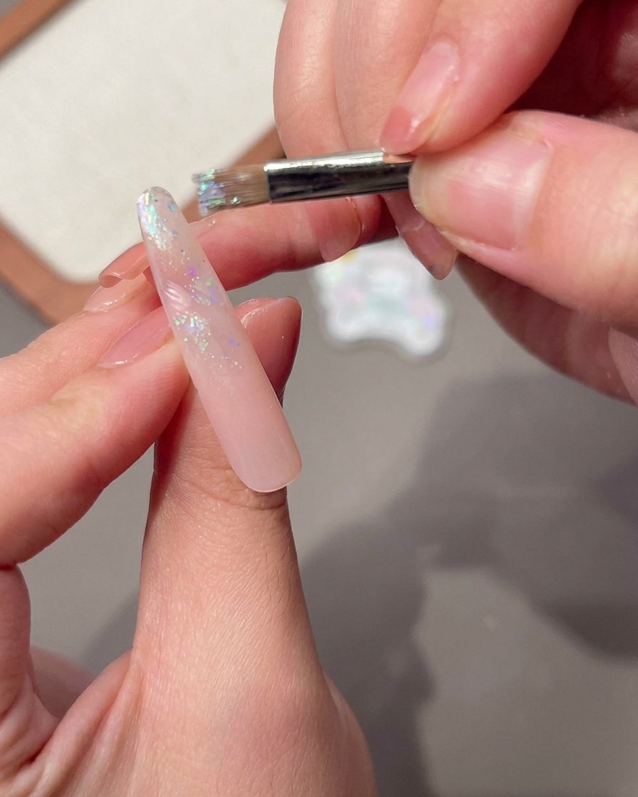 A nail brush applying a wet layer of base gel over the surface of a long pink nail.
