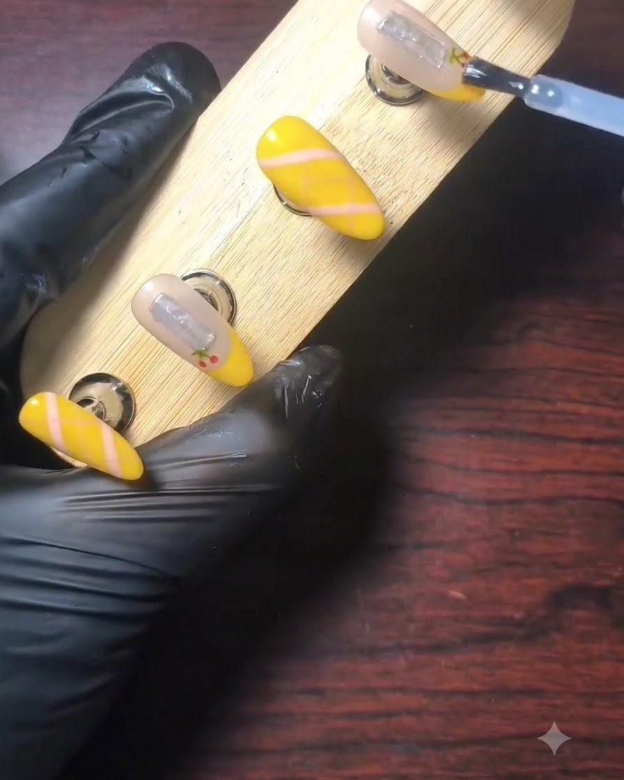 A gloved hand using a brush to apply clear shiny top coat over a decorated yellow French tip nail on a wooden display stand.
