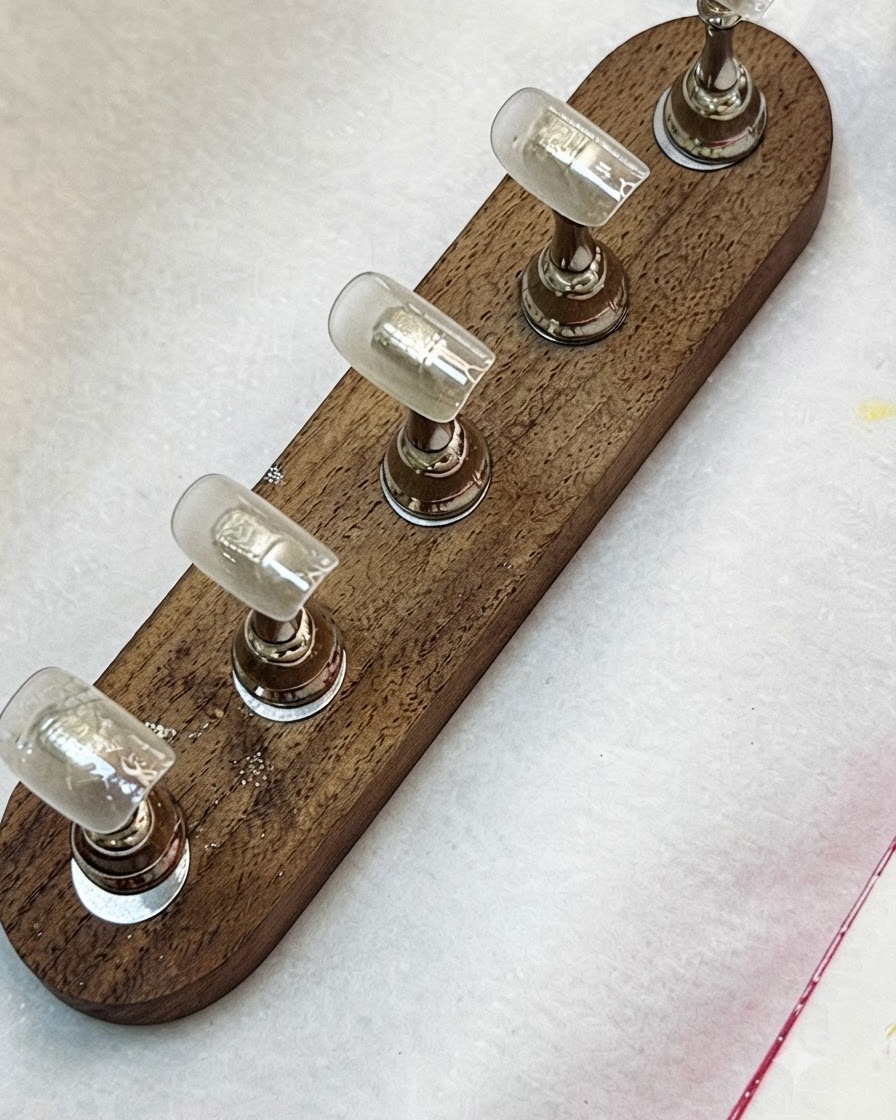 A brush applying clear base coat onto nail tips mounted on a wooden stand.