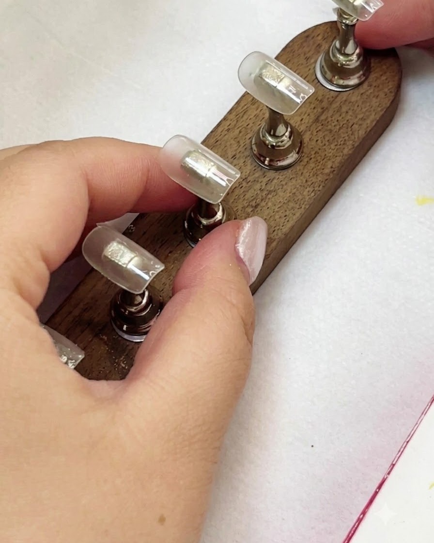 A hand holding a nail tip while applying a milky yellow gel color.