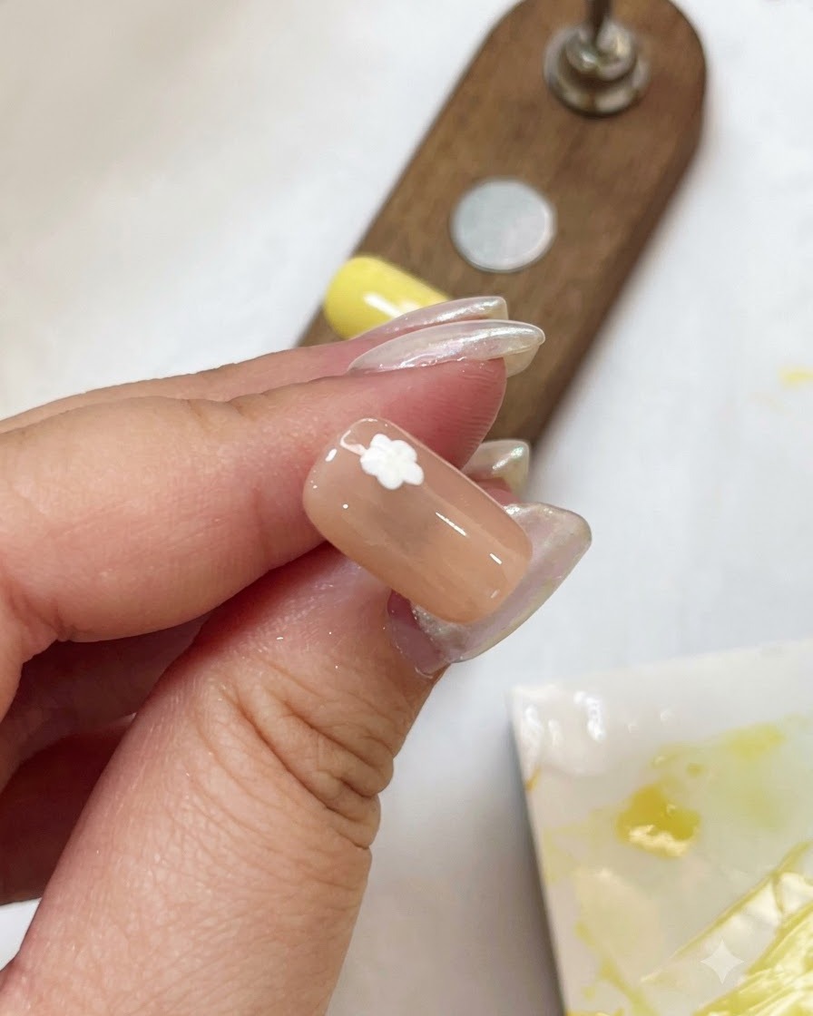 A close-up of a nude nail with a white flower petal design being cured using a small purple pen light.