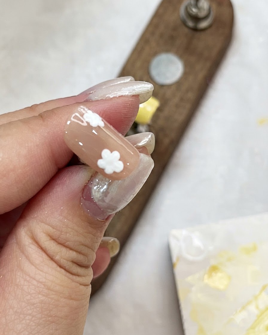 A hand holding a nude-colored nail tip while adding white dot petals to create flowers.
