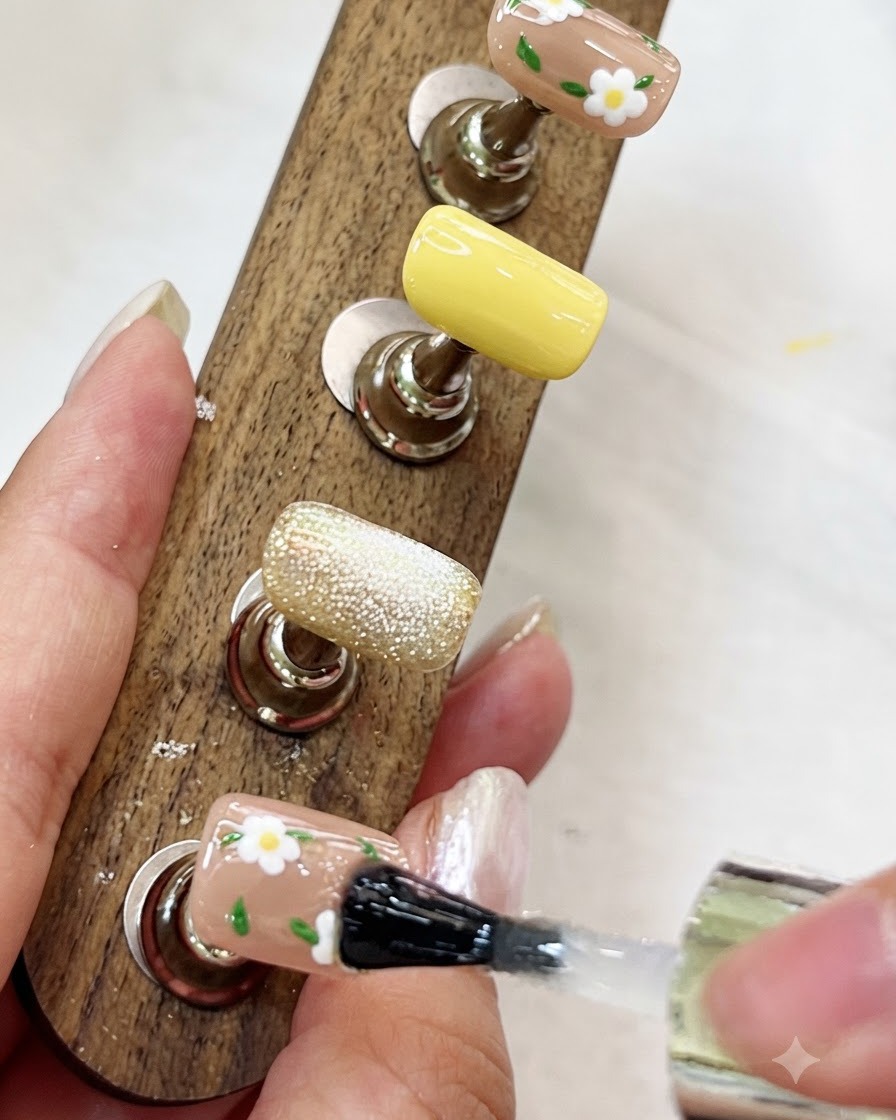 A brush applying a clear glossy top coat over a floral-painted nail on a wooden display stand.