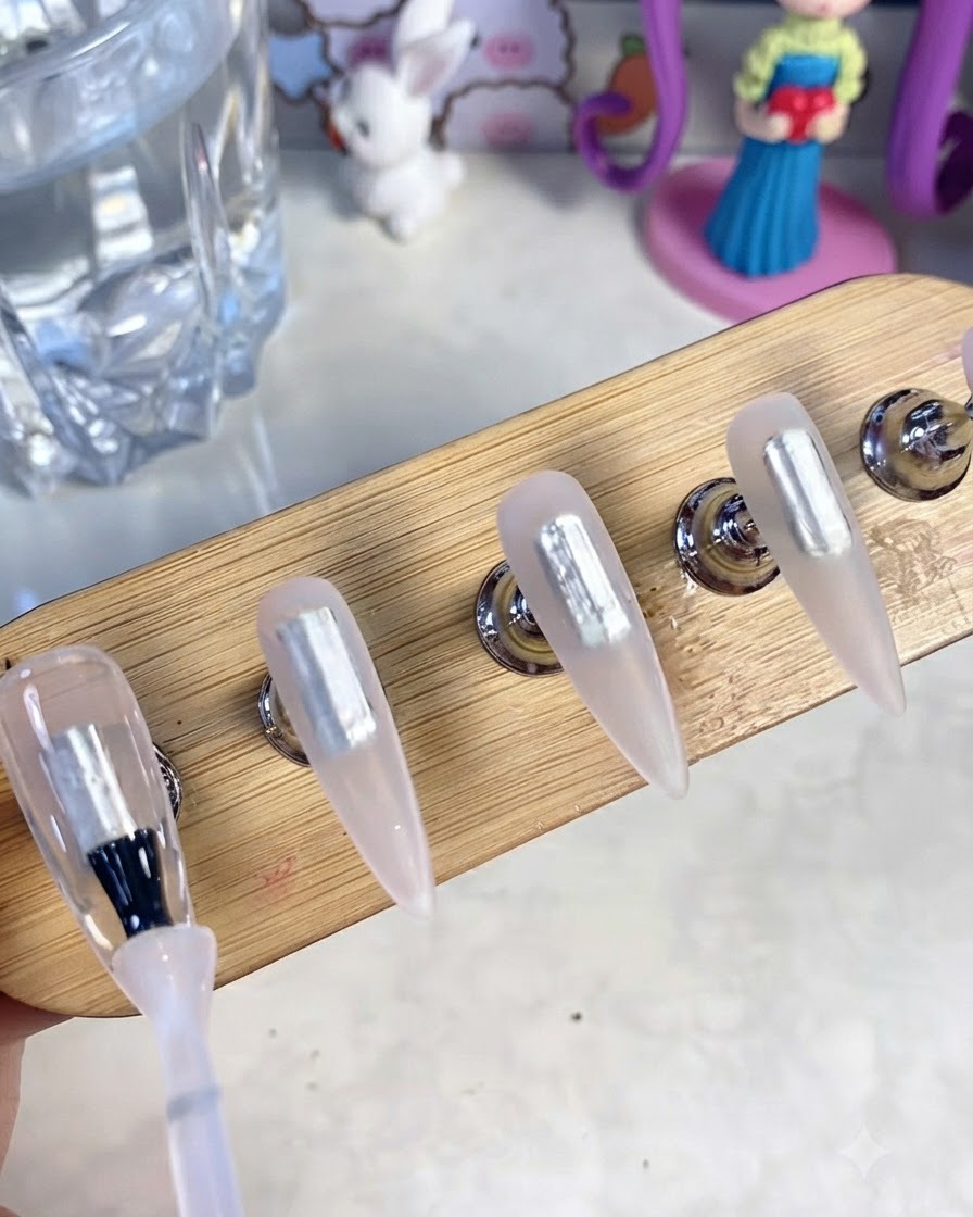 A close-up of a gel brush applying clear base gel onto blank artificial nail tips attached to a wooden display stand.