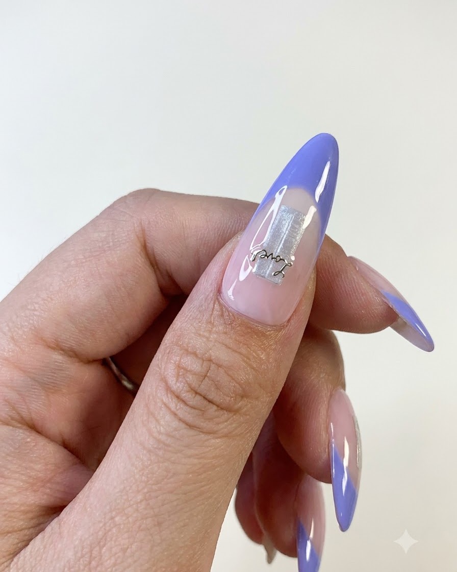 A close-up view of a thumb nail featuring a blue French tip, silver foil base element, and a delicate letter sticker.
