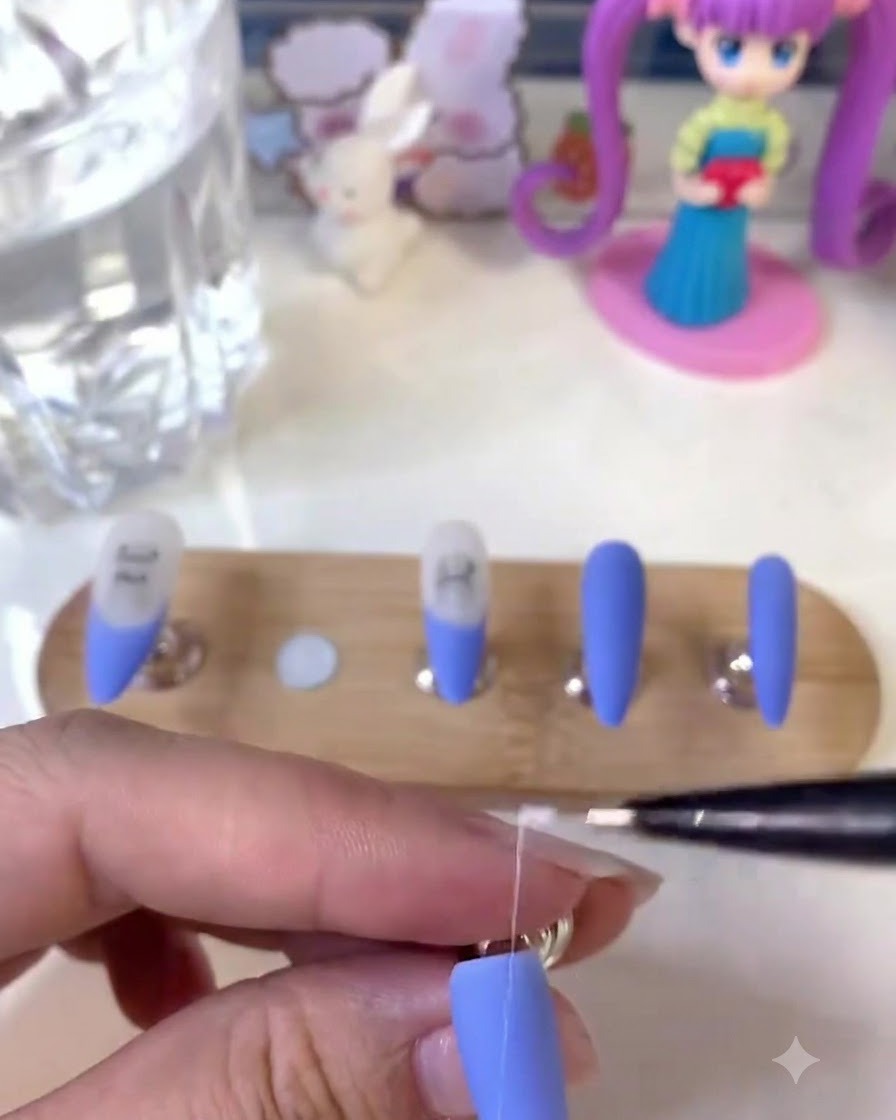 A nail artist holding a fine liner brush and drawing detailed white lines on a matte blue nail.