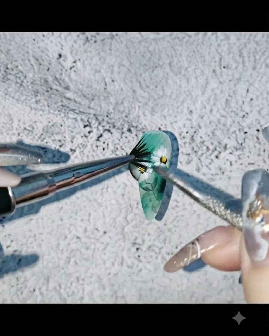 A nail artist flicking black watercolor paint onto a decorated green and floral nail tip.