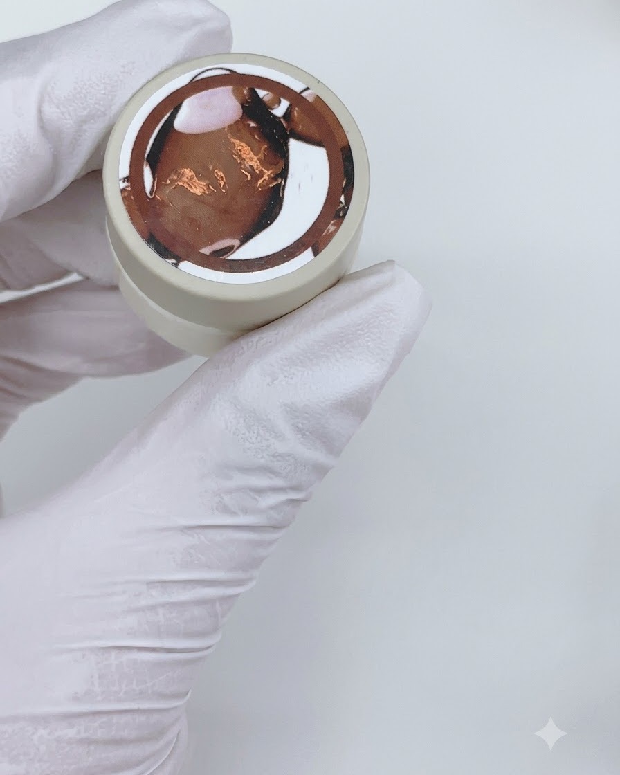A flat gel brush applying a coat of sheer brown gel polish onto a clear nail display stick.