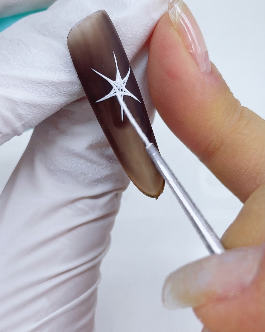 A close-up of a fine detail brush connecting the points and filling the center sections of a white starburst design over a matte brown gel base.