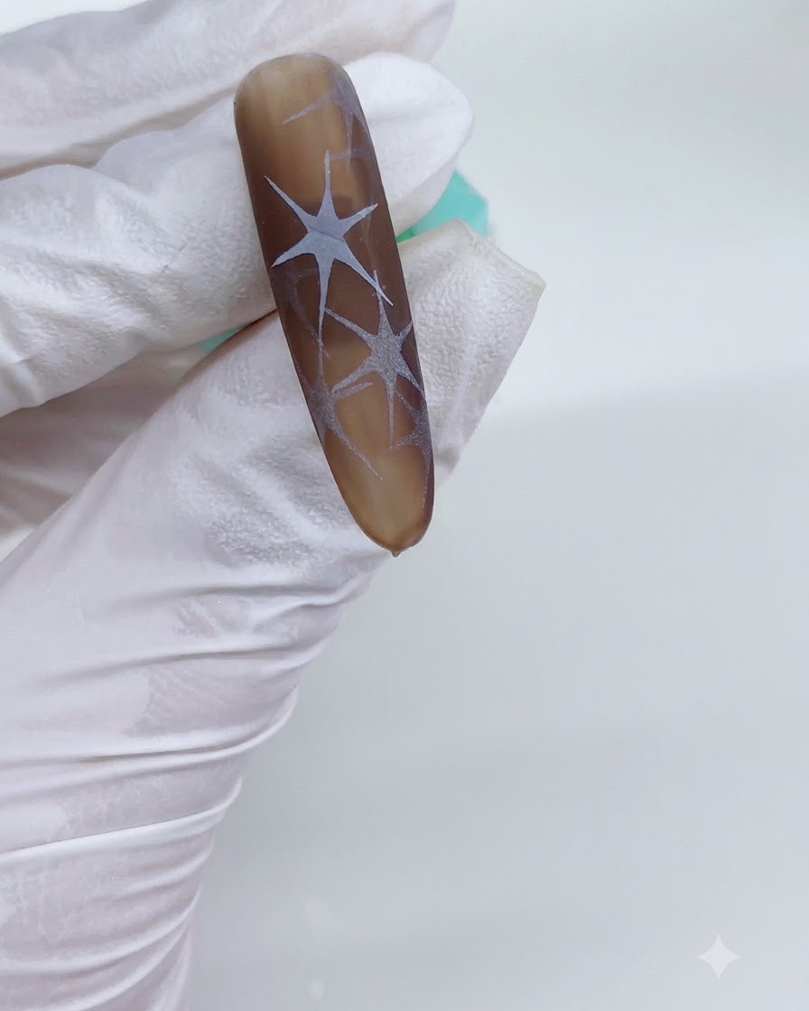A gloved hand holding a brown-toned nail under a UV lamp with cured white star patterns visible.