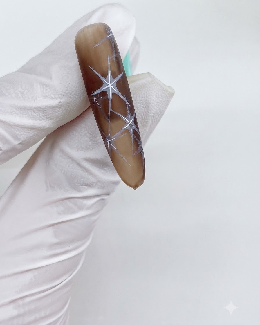 A close-up of a brown gradient nail with intricate white star patterns being cured.