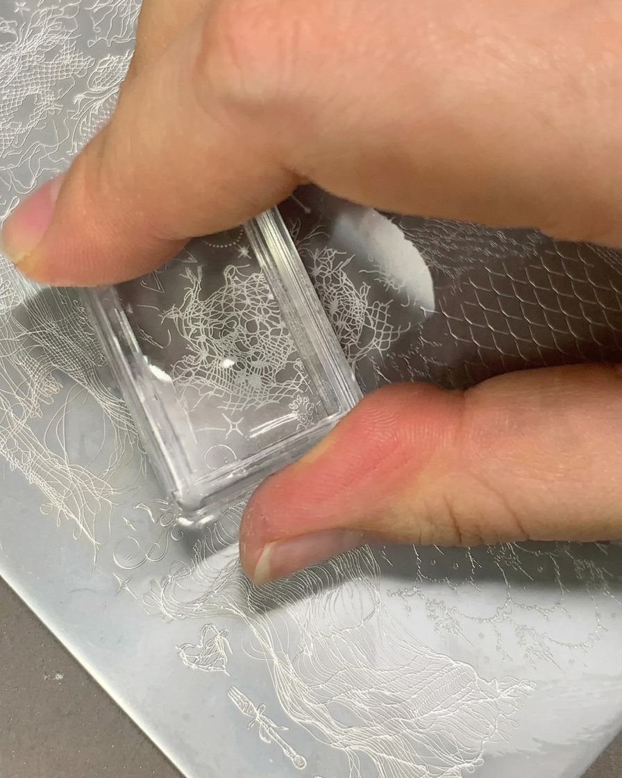 A clear stamper transferring a white lace pattern onto a clear nail tip.