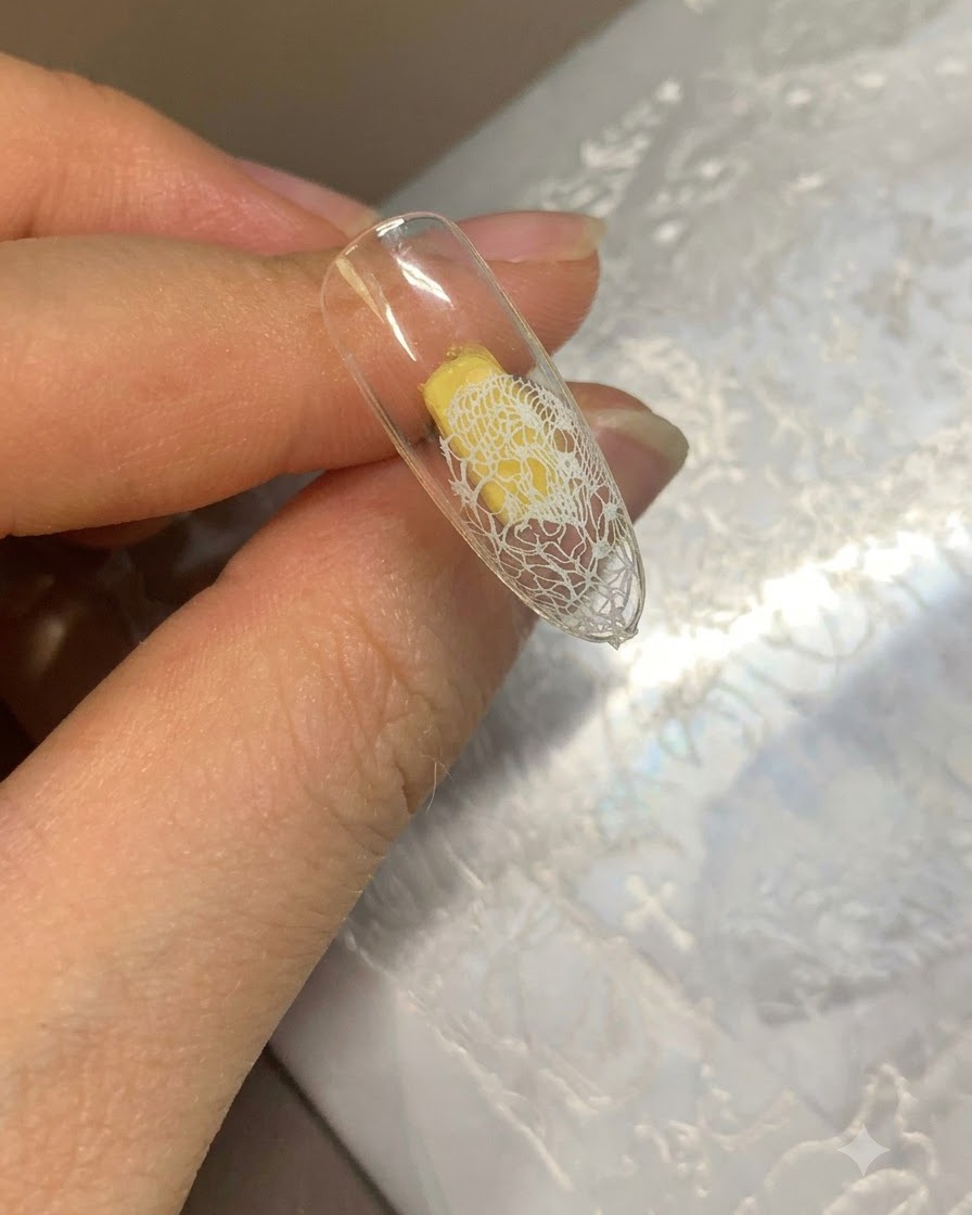 A clear nail tip showing a transferred white lace pattern at the end and a beige gel base applied to the rest.